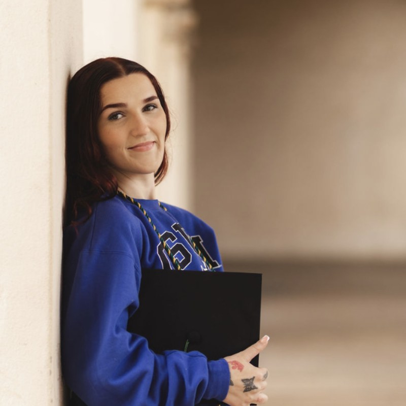 Kaitlyn J. Stowinsky leaning against a stone wall in a Cal State San Marcos sweatshirt, holding her graduation cap