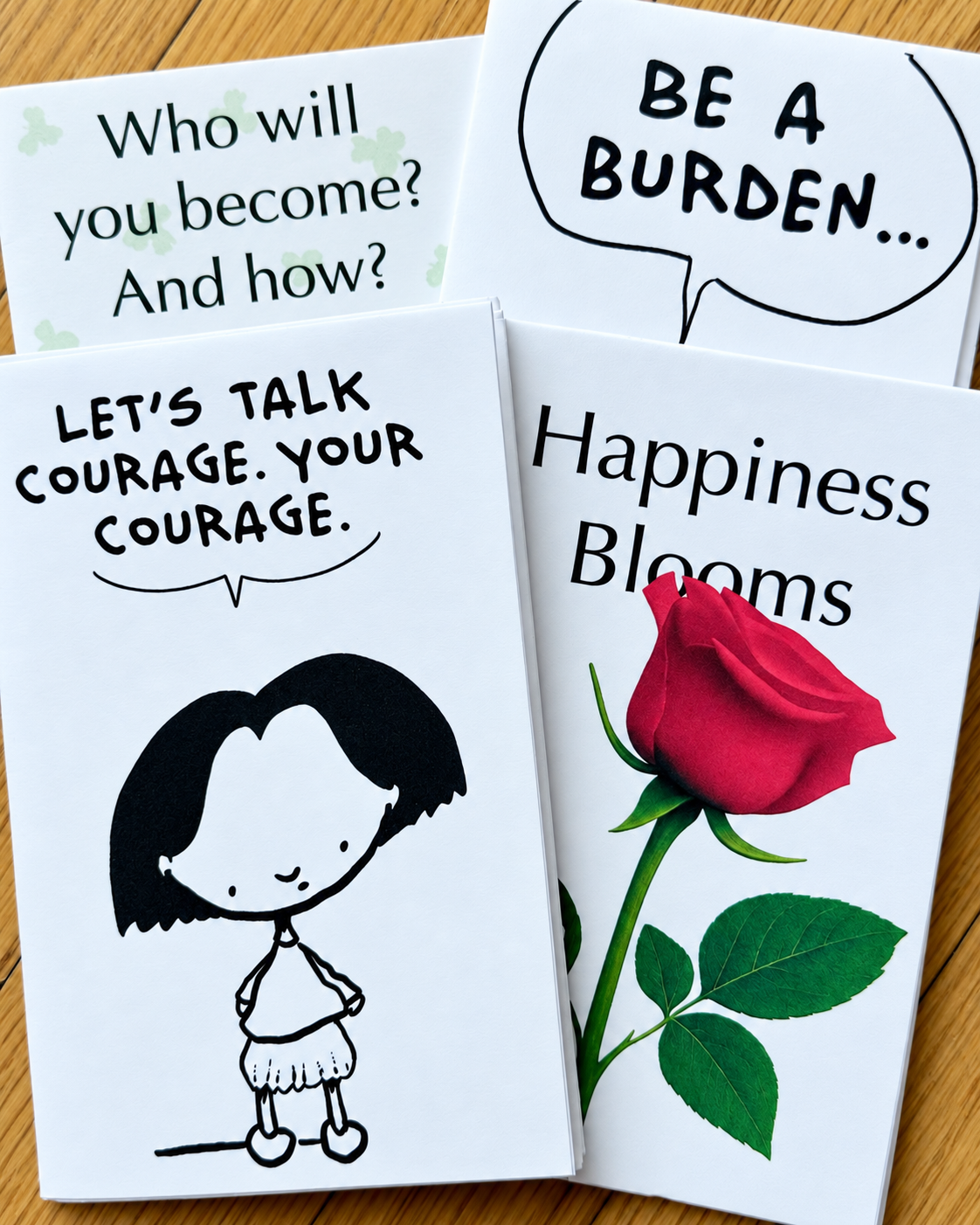 Four NextUp mini zines on a wooden table: Who will you become? And how?, Be a burden…, Let's talk courage. Your courage., and Happiness Blooms.